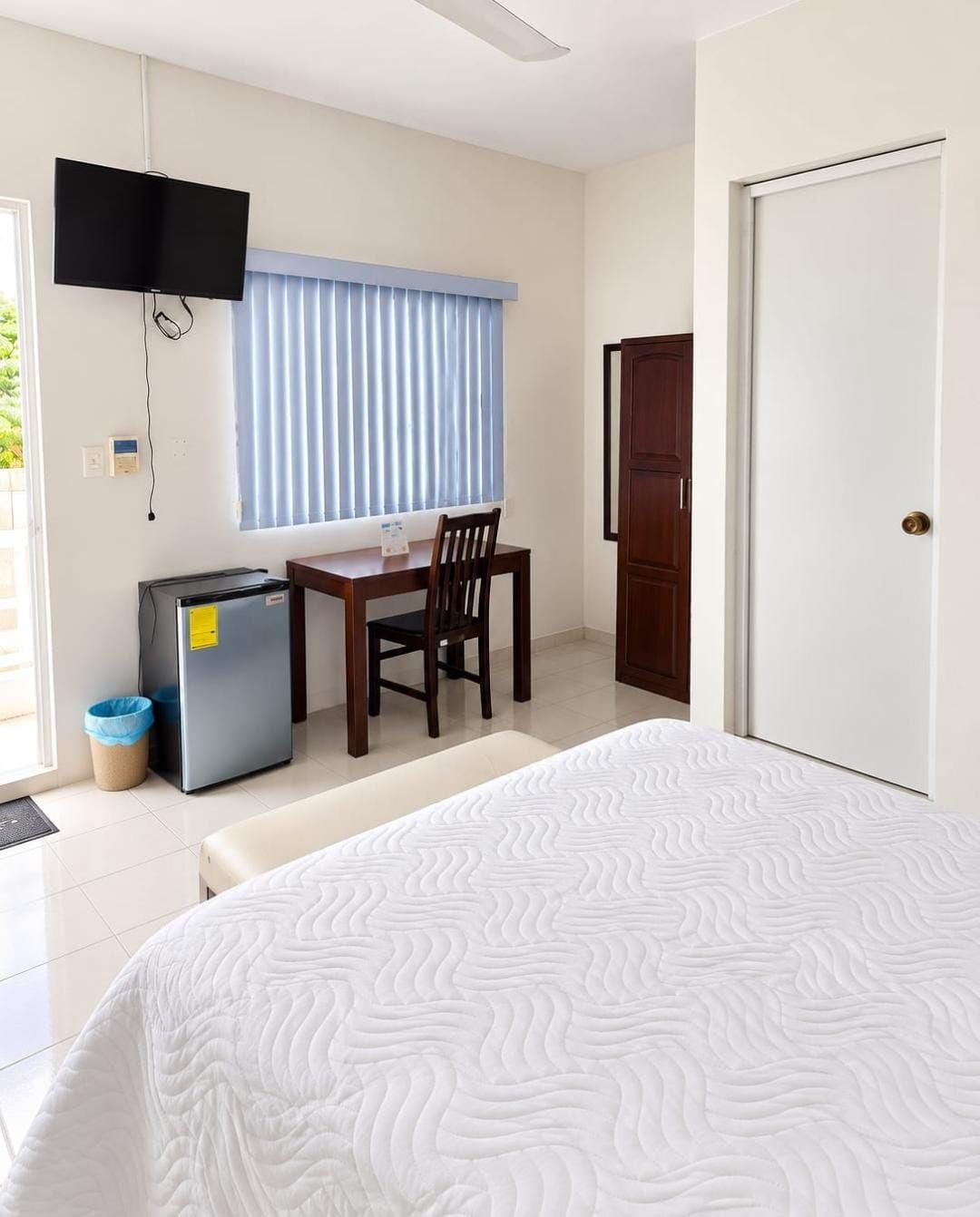 Un dormitorio con cama, escritorio con silla, televisión montada en la pared y una puerta.