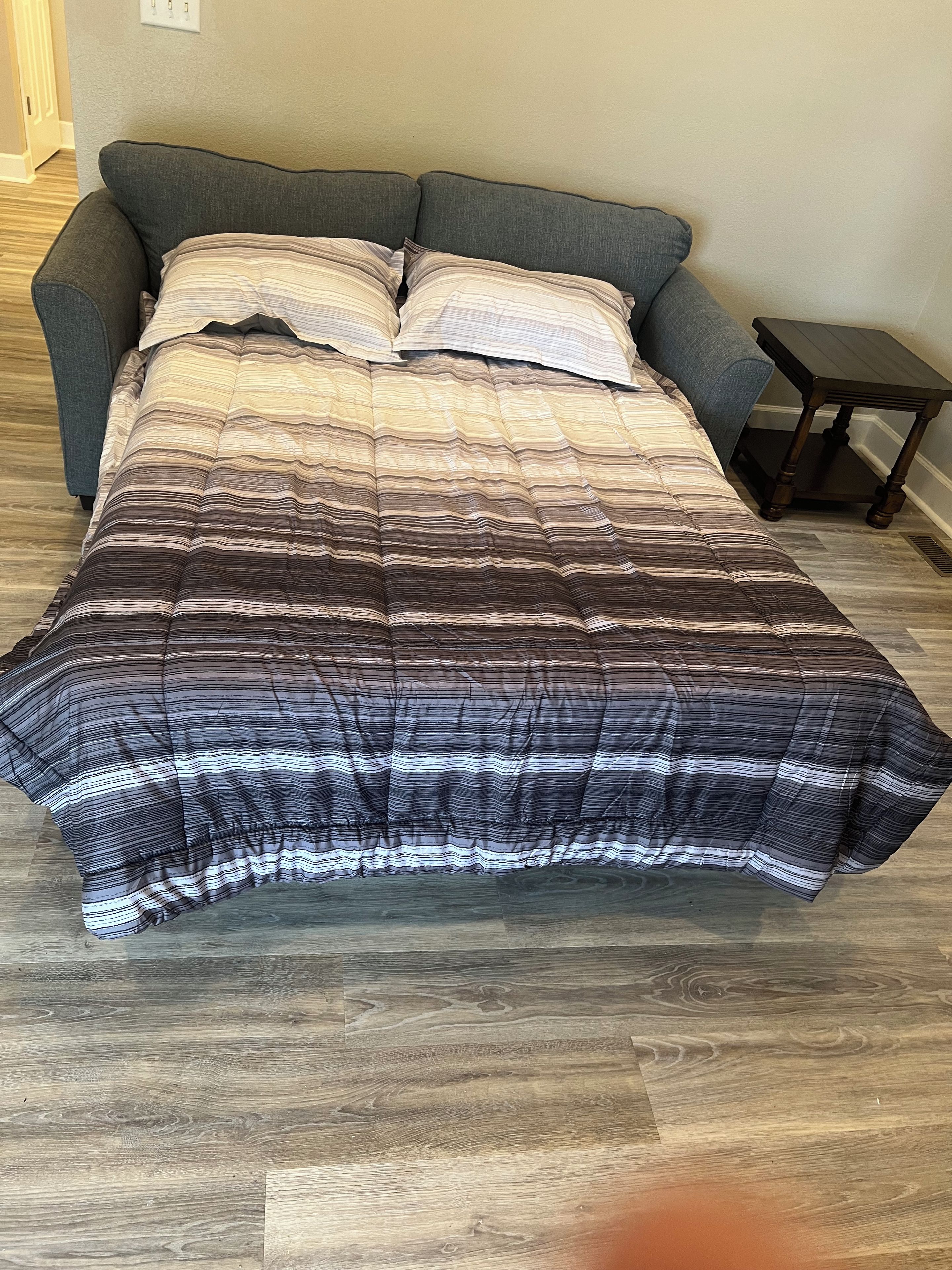 A bed with a striped comforter, a gray sofa, and a small wooden nightstand.