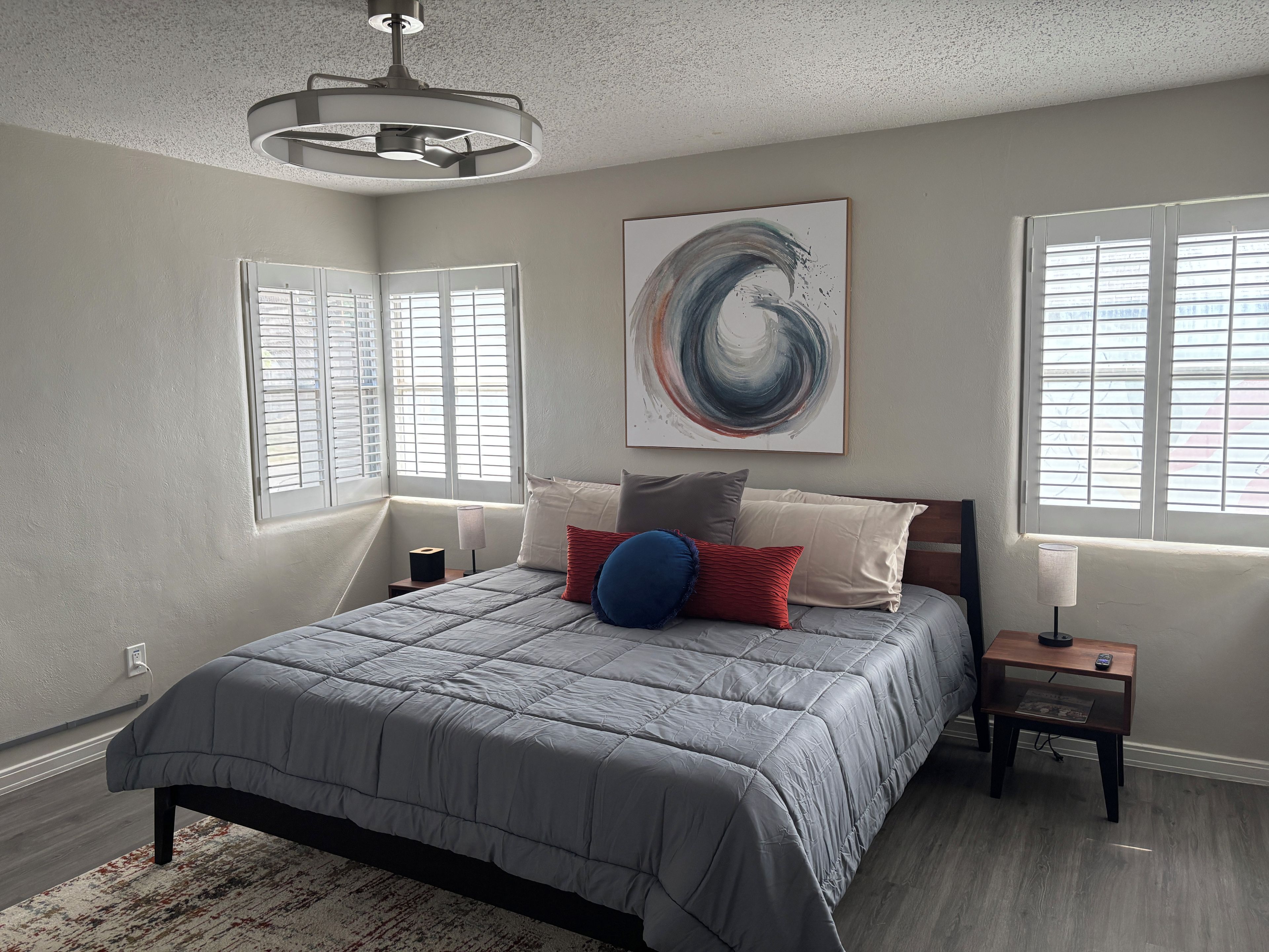 A bedroom with a bed, a nightstand, a lamp, a ceiling fan, and a window with blinds.