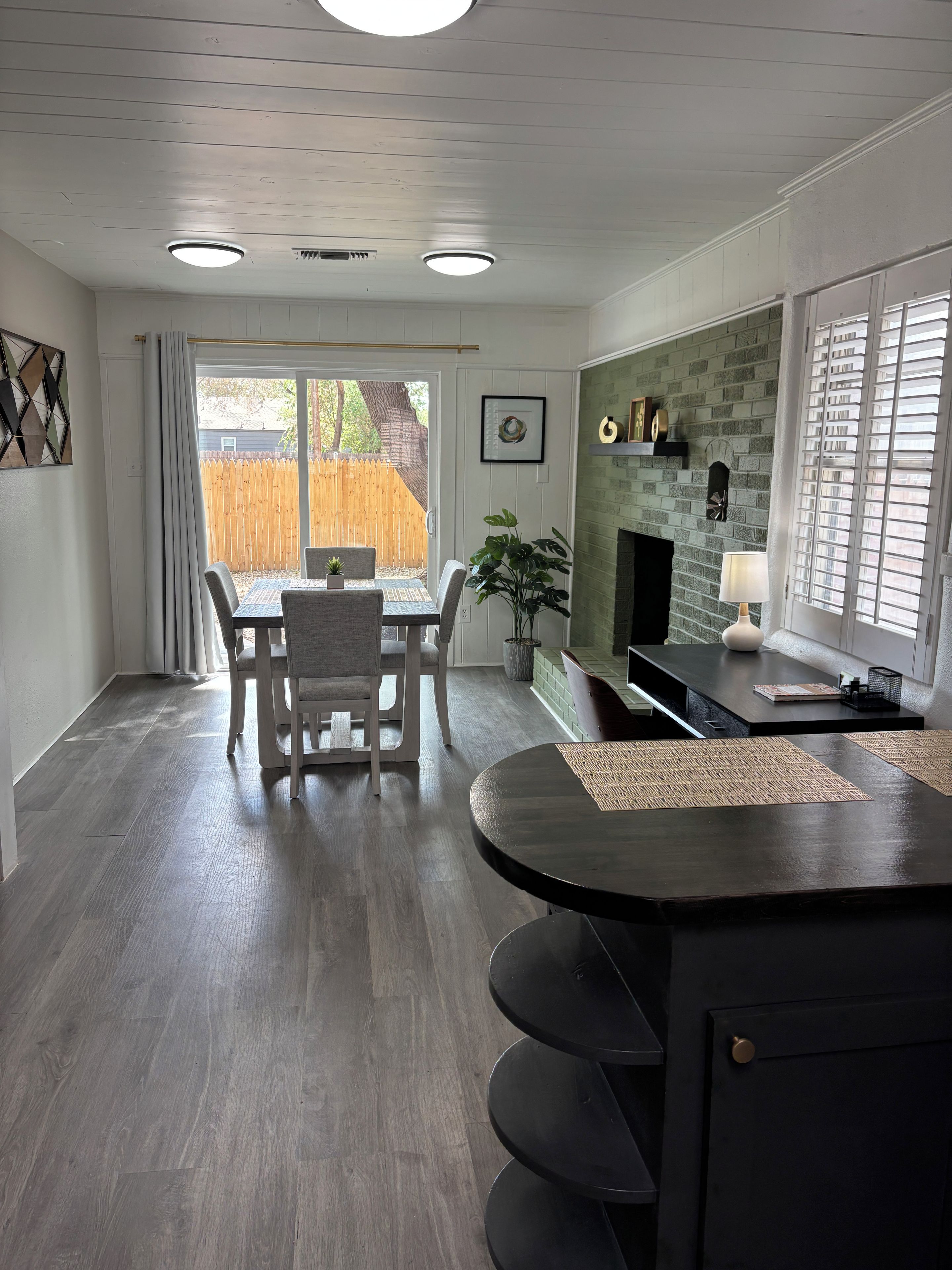 A modern kitchen with a dining area, featuring a wooden floor, a fireplace, and a view of the backyard.