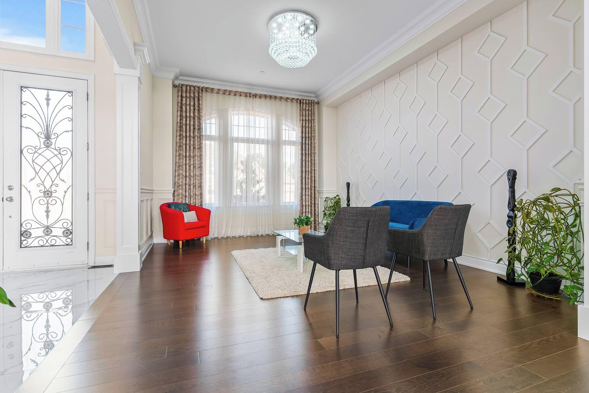 A modern living room with a wooden floor, a red armchair, a grey sofa, a chandelier, and a geometric wall design.
