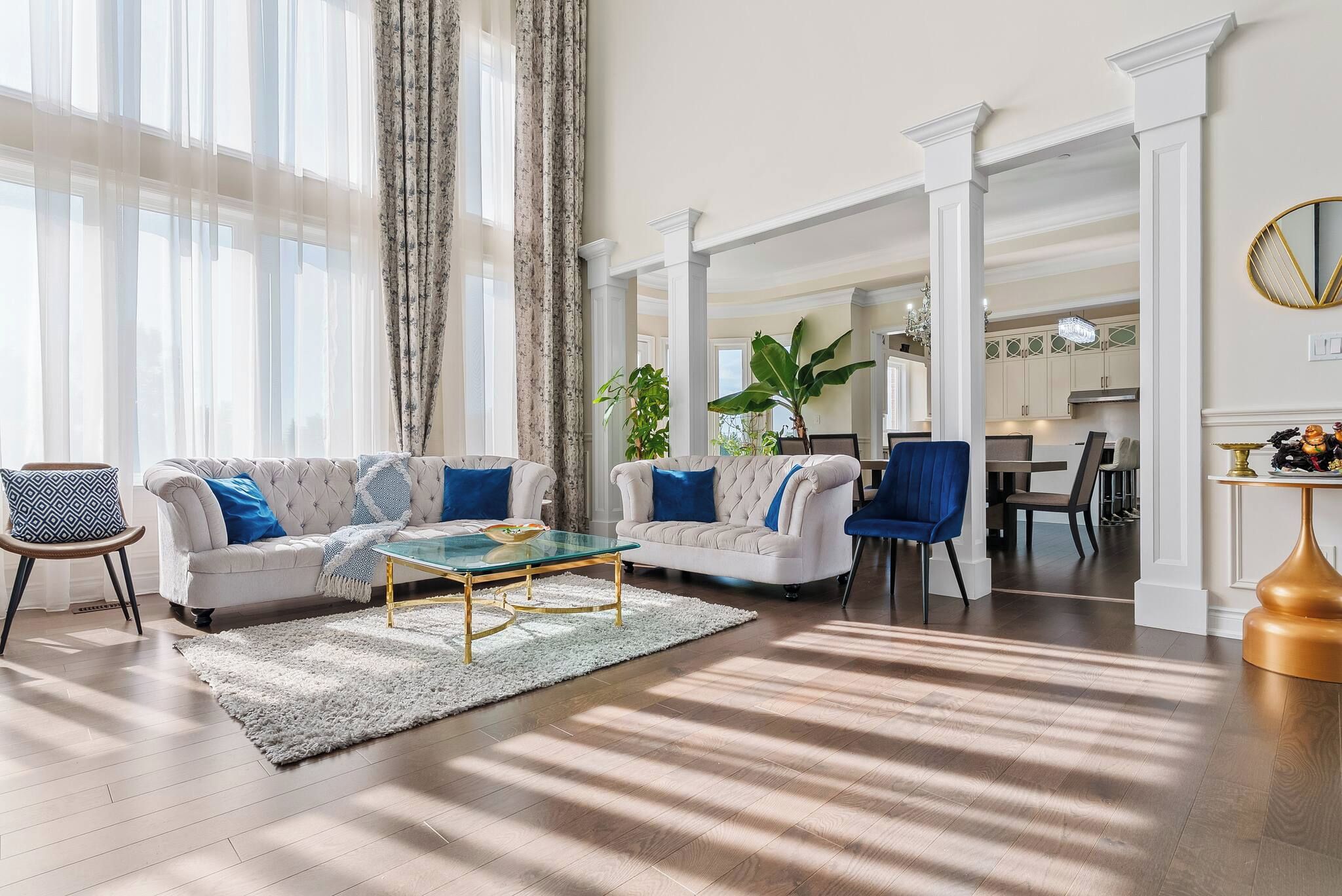 Amplia sala de estar con un ventanal grande, un sofá blanco, un sillón azul, una mesa de centro de vidrio y una zona de comedor al fondo.