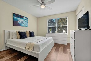 Une chambre à coucher avec un lit, un ventilateur de plafond, une fenêtre avec des stores et un téléviseur posé sur un meuble.