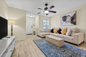 Ein Wohnzimmer mit einem beigefarbenen Ecksofa, einem blauen Teppich mit Muster, einem Deckenventilator und einem Flachbildfernseher.