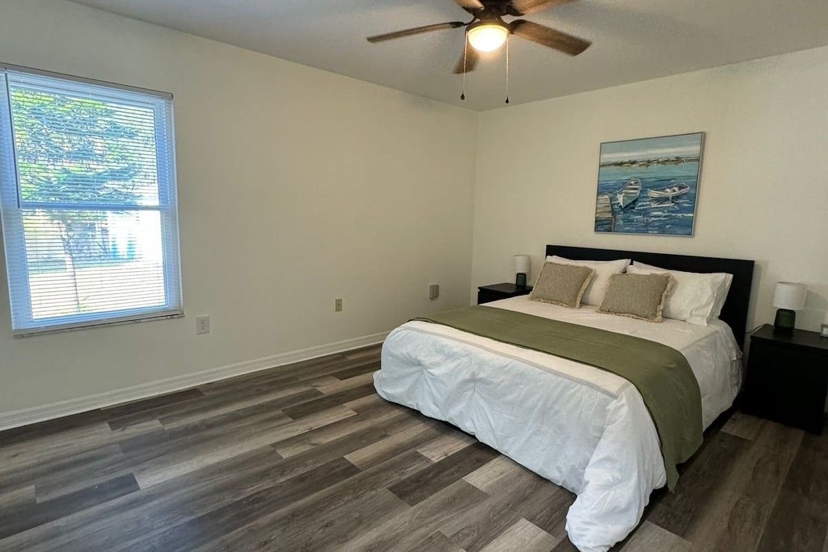 Une chambre à coucher avec un lit, un ventilateur de plafond, une fenêtre avec des stores et un tableau accroché au mur.