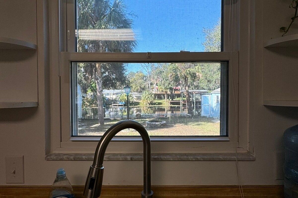 A window with a metal faucet in the foreground, a clear view of a backyard with a fence and trees.