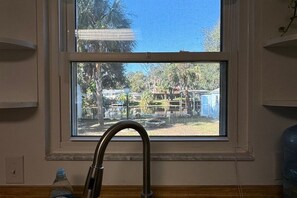 Una ventana con un grifo metálico al frente, vista despejada a un patio trasero con cerco y árboles.