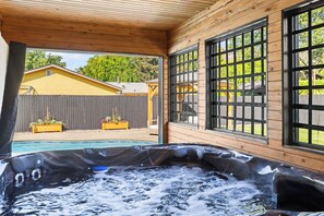 Ett bubbelbad med en trädeck och stora fönster, med utsikt över en trädgård med staket och en gul byggnad.