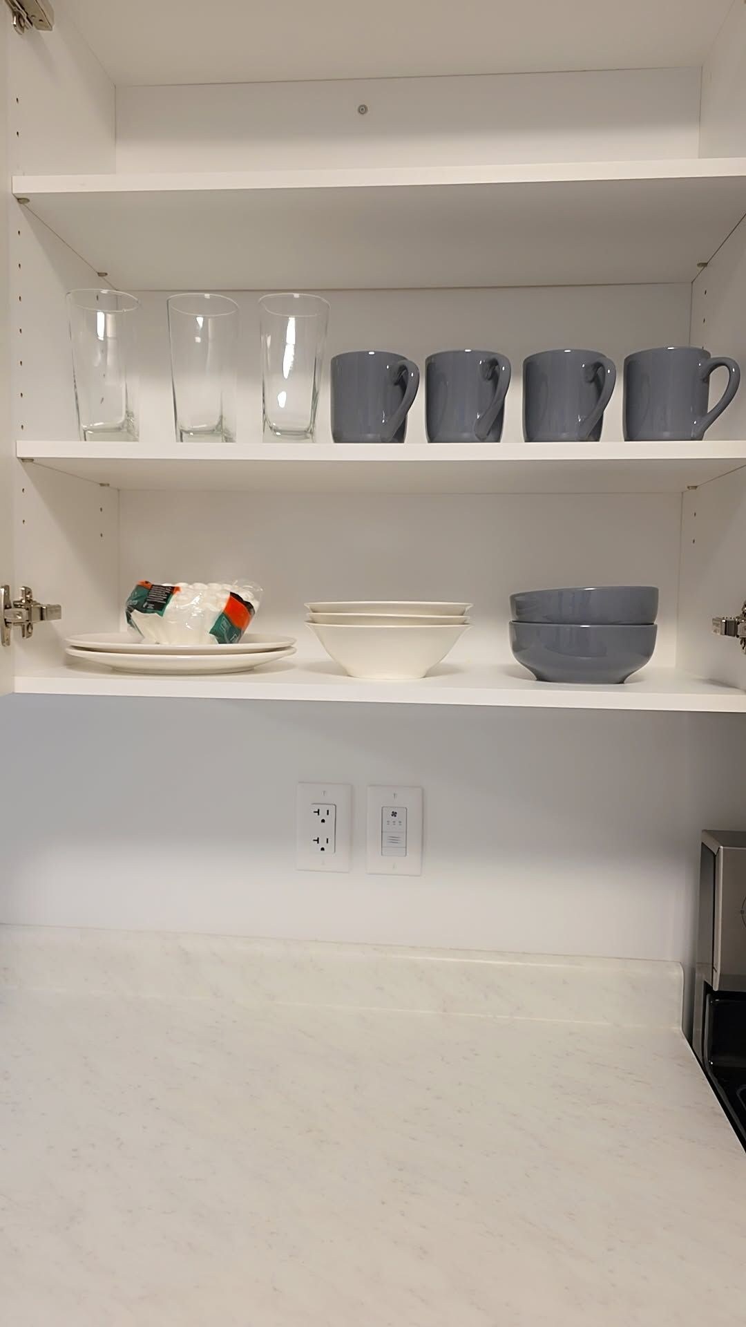 A kitchen with white cabinets, a countertop, and a shelf with various kitchenware including cups and bowls.