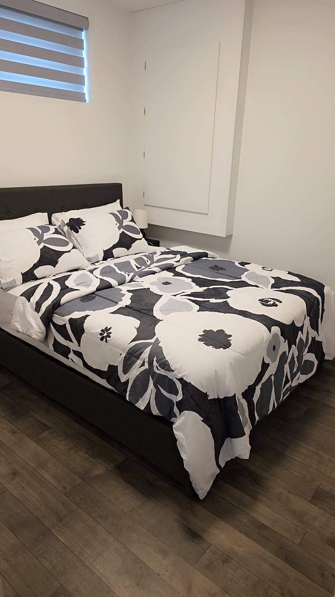 A bed with a floral-patterned bedspread, a dark headboard, and a window with blinds.