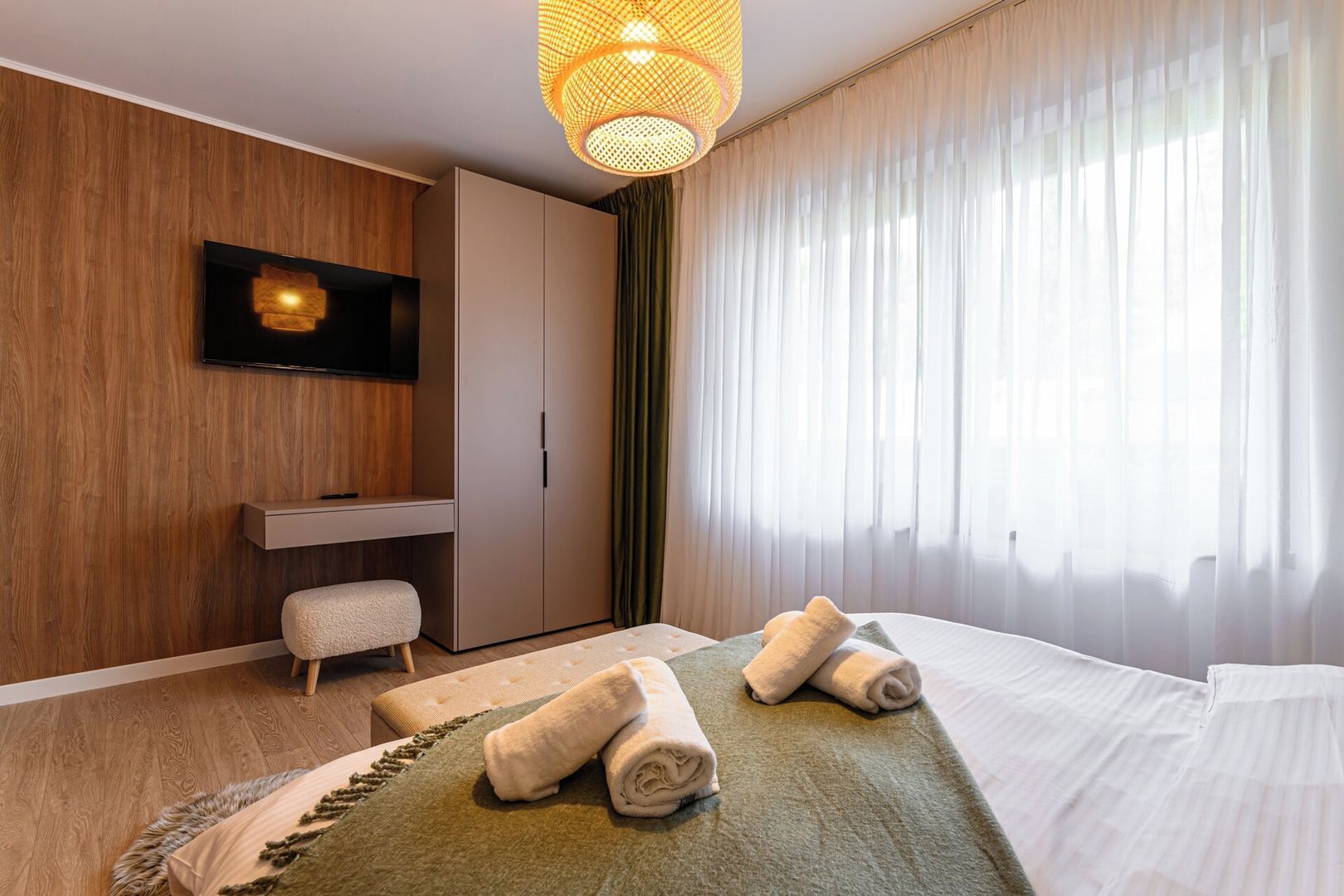Hotellihuone, jossa on sänky, taulutelevisio, vaatekaappi ja suuri ikkuna, jossa on läpikuultavat verhot.