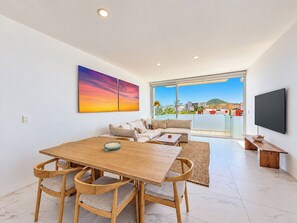 A modern living room with a wooden dining table, cushioned chairs, a sofa, and a large window offering a view of buildings and greenery. - Los Cabos Chic Stay Walk to Beach & Nightlife (Cabo San Lucas)