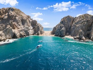 A coastal scene with rocky cliffs, clear turquoise waters, and a boat in the foreground. - Los Cabos Chic Stay Walk to Beach & Nightlife (Cabo San Lucas)