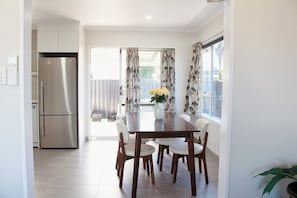 A dining area with a wooden table and chairs, a refrigerator, and a window with curtains. - Sunny modern 3BR home in Napier - Near supermarket, dining and parks (Napier)