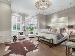 A bedroom with a large bed, a chandelier, a cowhide rug, and a view of buildings through large windows. - The Chelsea Town House (London)