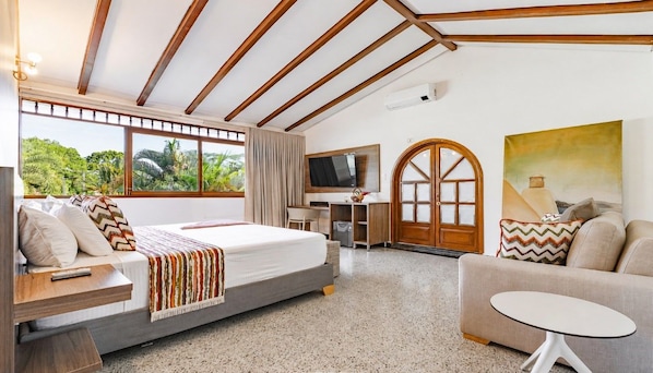 A hotel room with a bed, a sofa, a desk with a TV, a painting, and a view of greenery.