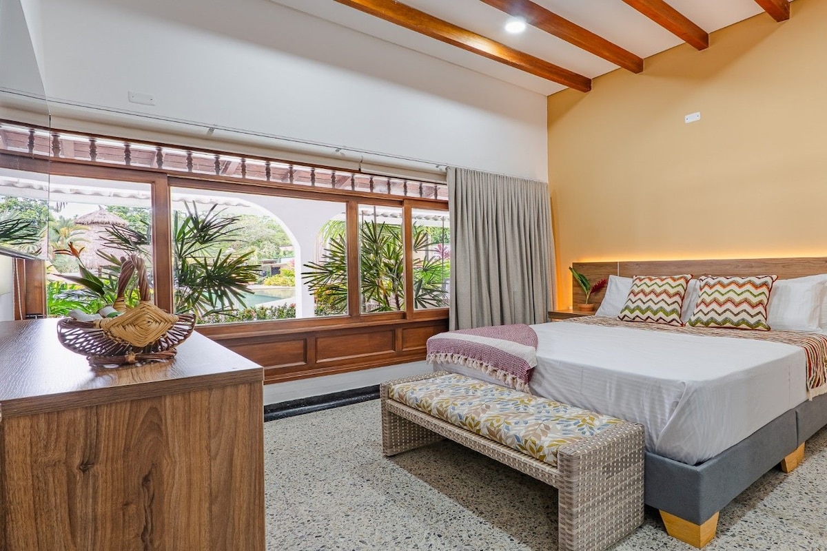 A bedroom with a bed, bedside table, bench, and a view of a garden through large windows.