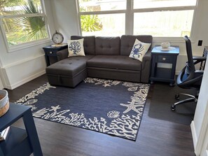 Ein Wohnzimmer mit einem Ecksofa, einem kleinen Hocker, einem Teppich mit Meeresmotiv, einer Uhr auf einem Beistelltisch und Blick auf einen Holzzaun durch ein Fenster.