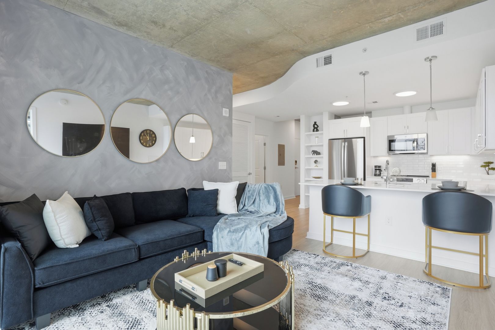 Un salon moderne avec un canapé bleu foncé, une table basse ronde et un coin cuisine doté de meubles blancs et d’un réfrigérateur en acier inoxydable.