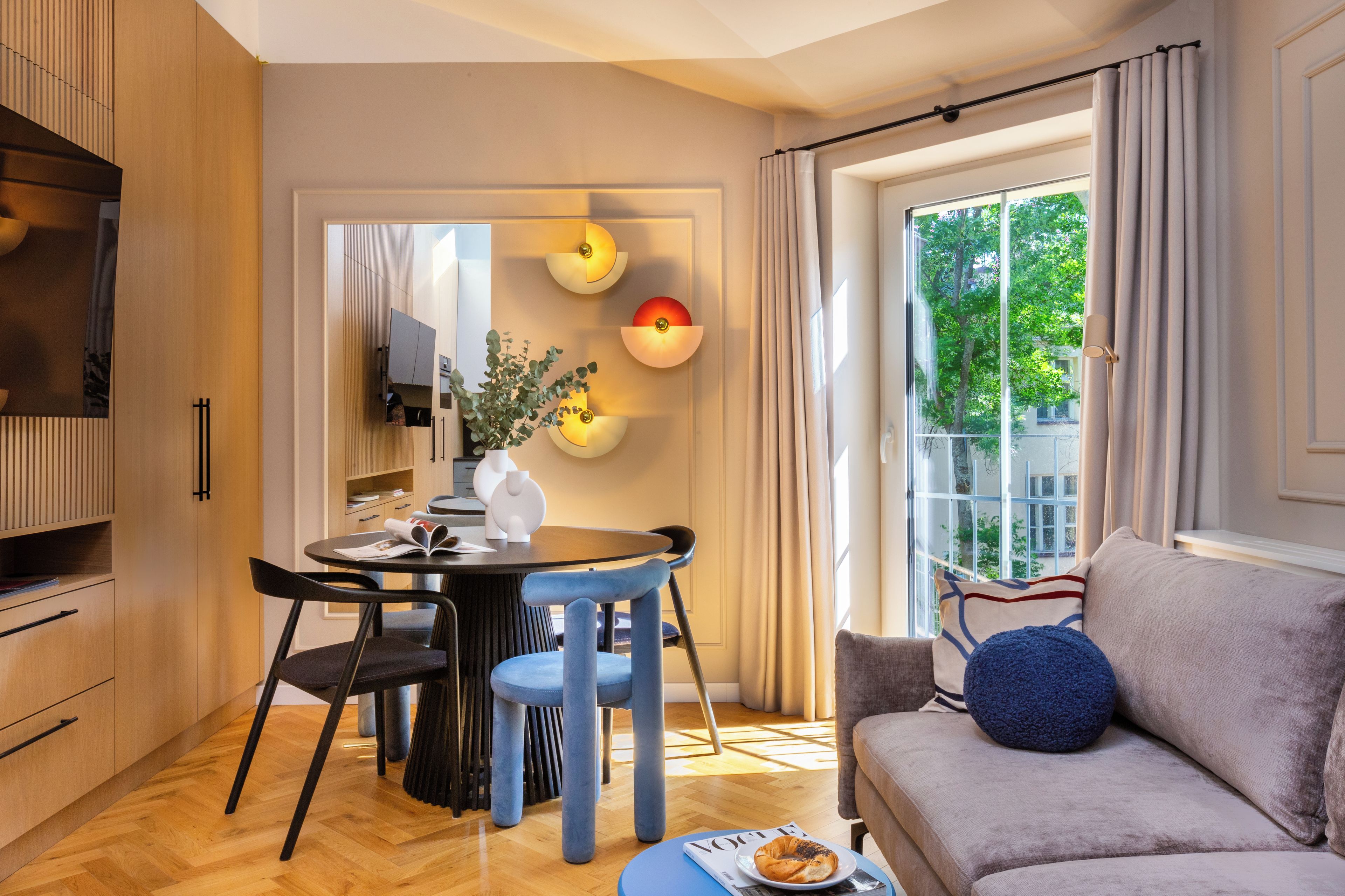 A modern living room with a sofa, a dining table with chairs, a TV, and a large window with curtains.