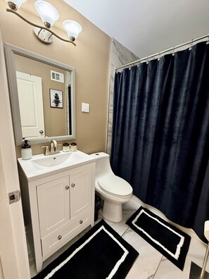A bathroom with a white vanity, a toilet, a dark blue shower curtain, and two black and white rugs. - Peaceful 3-BR House in Vibrant Memphis (Memphis)