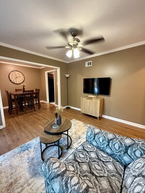 A living room with a ceiling fan, a dining area with a clock, a television, and a patterned sofa. - Peaceful 3-BR House in Vibrant Memphis (Memphis)