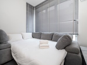 A modern bedroom with a sectional sofa, a bed with a white comforter, and large windows with blinds. - Piotrkowska Modern Apartment (Łódź)