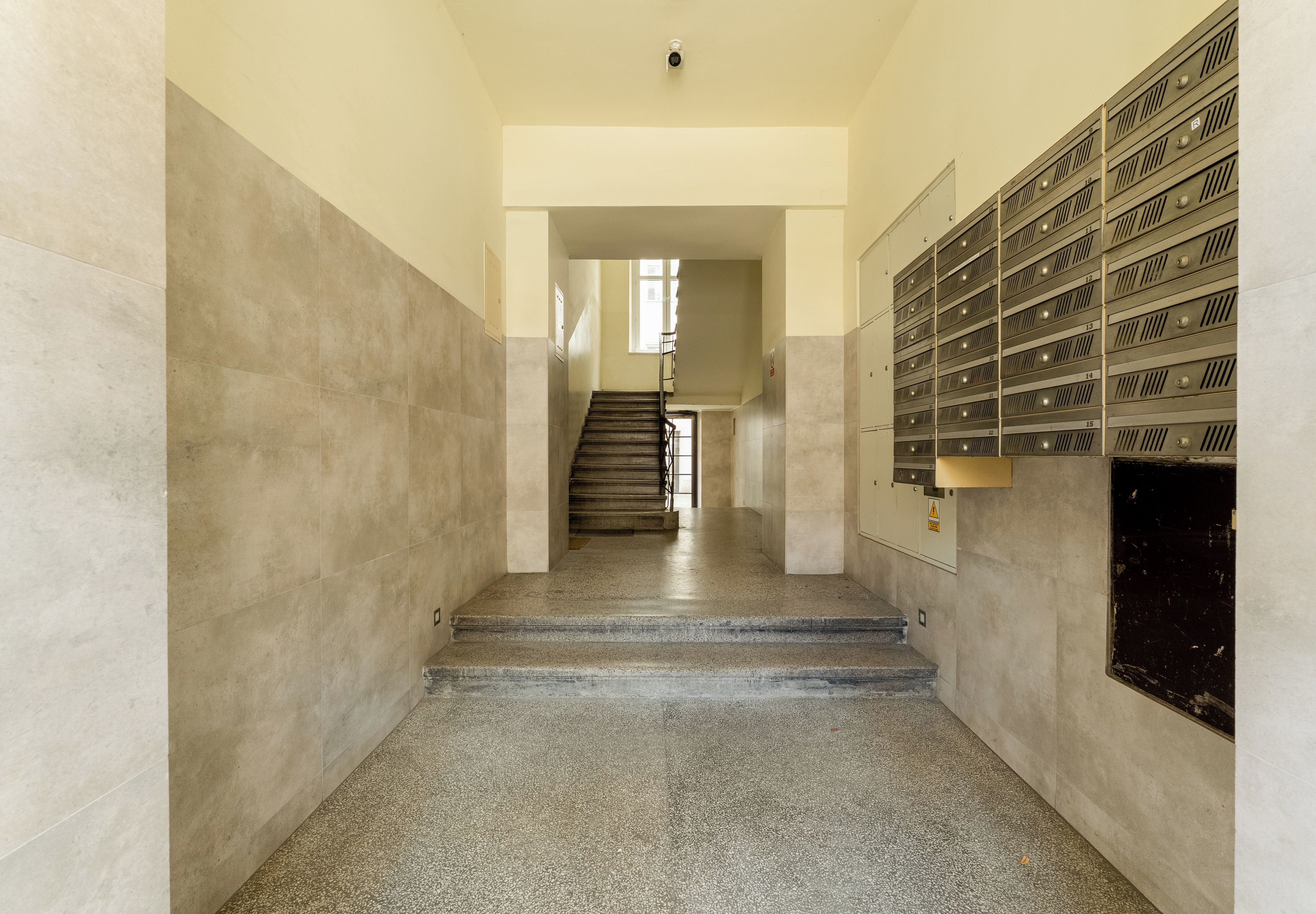 A hallway with a staircase leading to a landing, a row of lockers on the right, and a door at the end of the hallway.