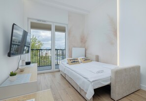 A modern bedroom with a bed, a TV, a balcony, and a view of the city. - Artyleryjska Superior Apartment (Kołobrzeg)