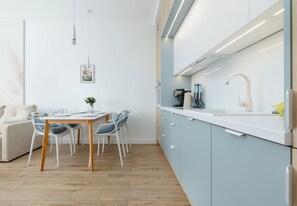 A modern kitchen with light blue cabinets, a white countertop, and a wooden dining table with chairs. - Artyleryjska Superior Apartment (Kołobrzeg)