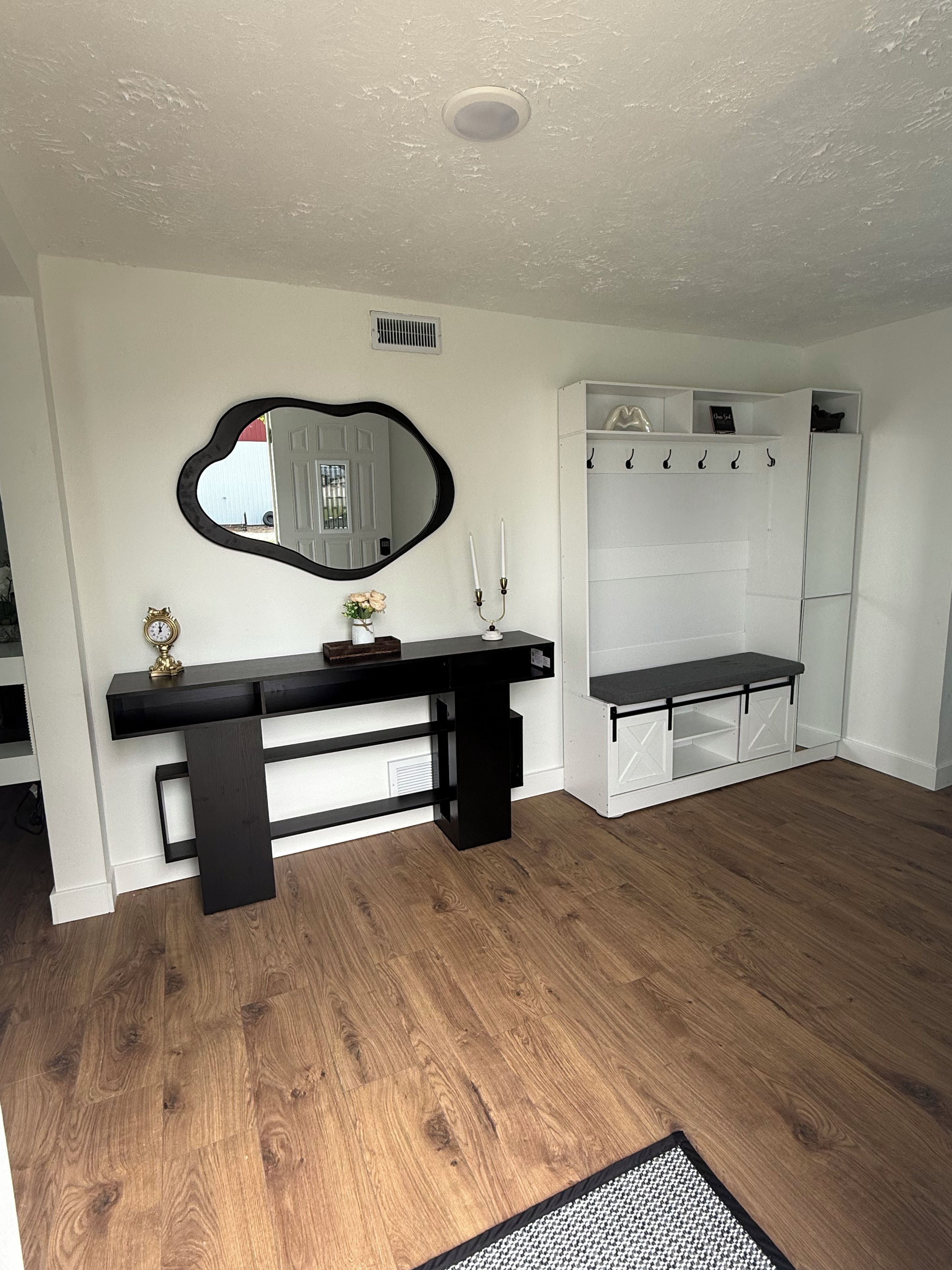 A room with a wooden floor, a black console table with a mirror, a white coat rack, and a bench.