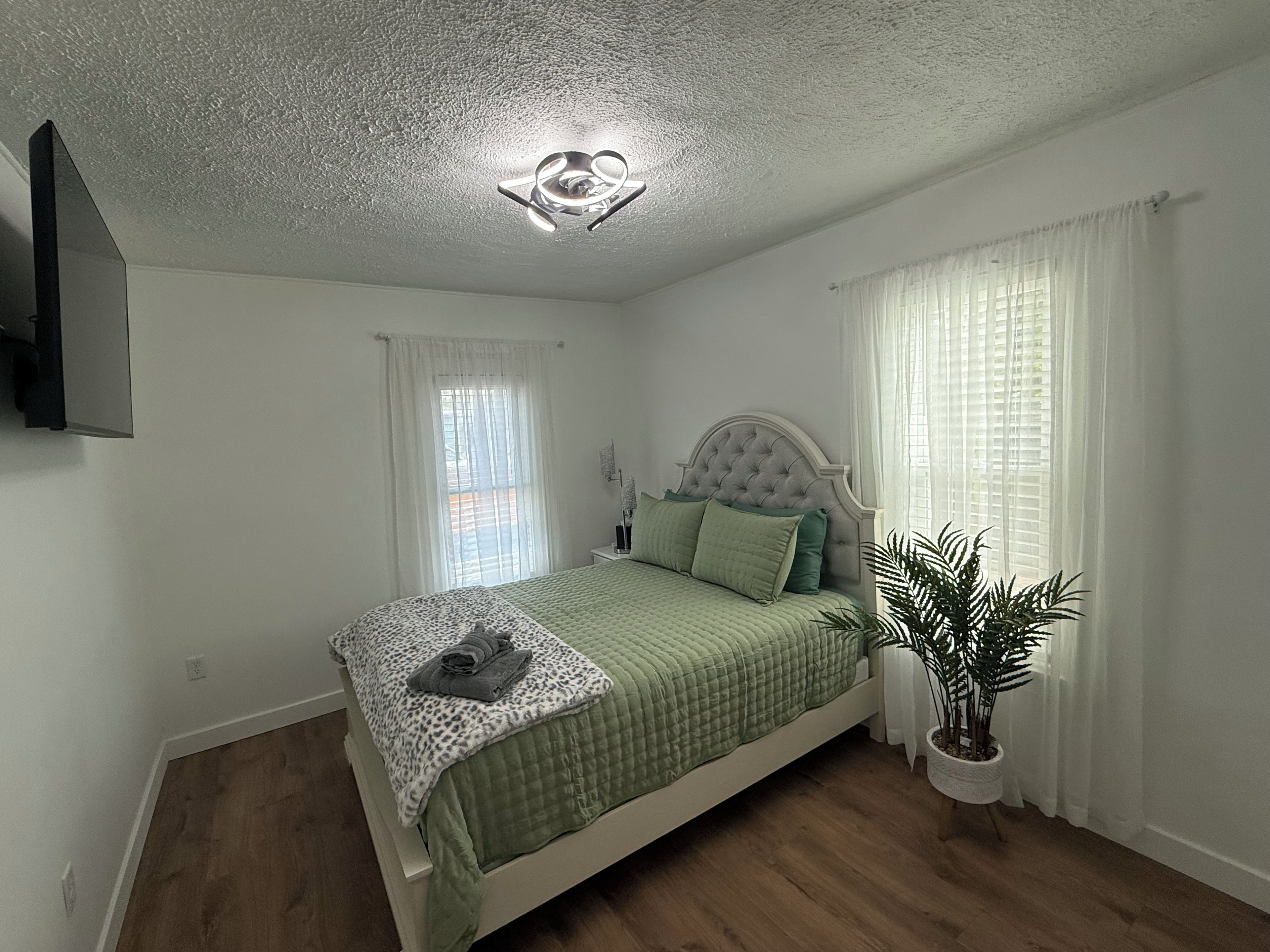 A bedroom with a bed, a ceiling light fixture, a potted plant, and a window with curtains.