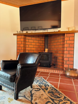 A living room with a brick fireplace, a leather armchair, a flat-screen TV mounted above the fireplace, and a patterned rug on the floor. - 4-bedroom house in Melides with an outdoor hot tub. (Melides)