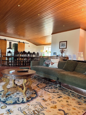 A cozy living room with a wooden ceiling, a green sofa, a patterned rug, and a dining area with wooden chairs and a table. - 4-bedroom house in Melides with an outdoor hot tub. (Melides)
