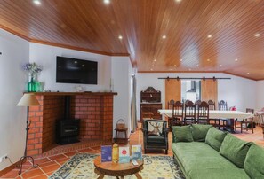A living room with a brick fireplace, a television mounted on the wall, a dining area with a table and chairs, and a green sofa. - 4-bedroom house in Melides with an outdoor hot tub. (Melides)