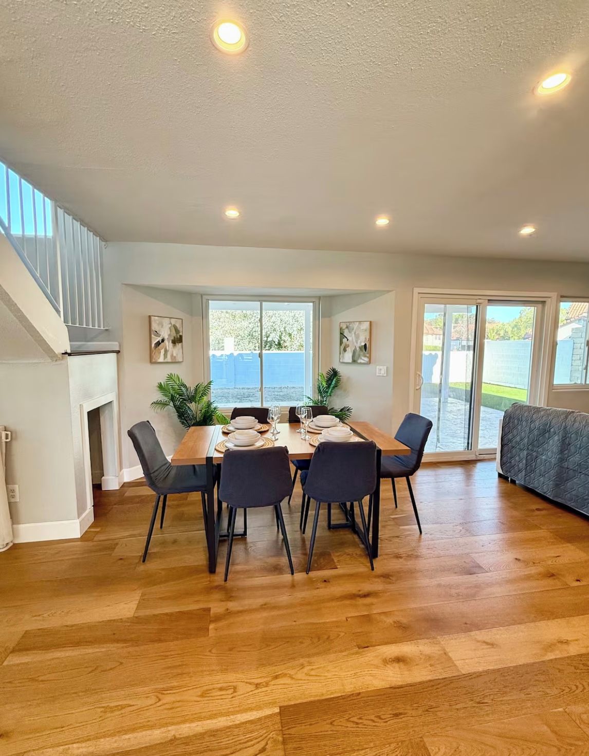 A dining area with a wooden table set for six, surrounded by chairs, with a view of the outdoors through large windows.