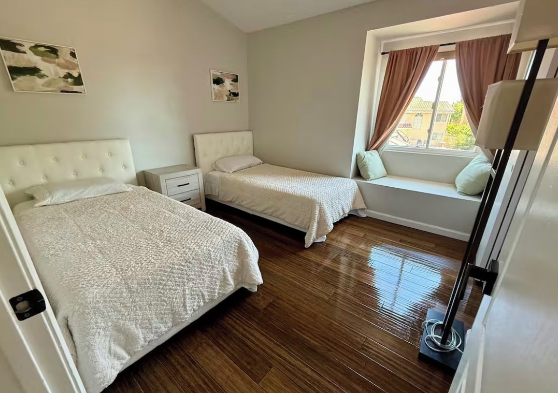 A bedroom with two beds, a window seat with curtains, and a bedside table with a lamp.
