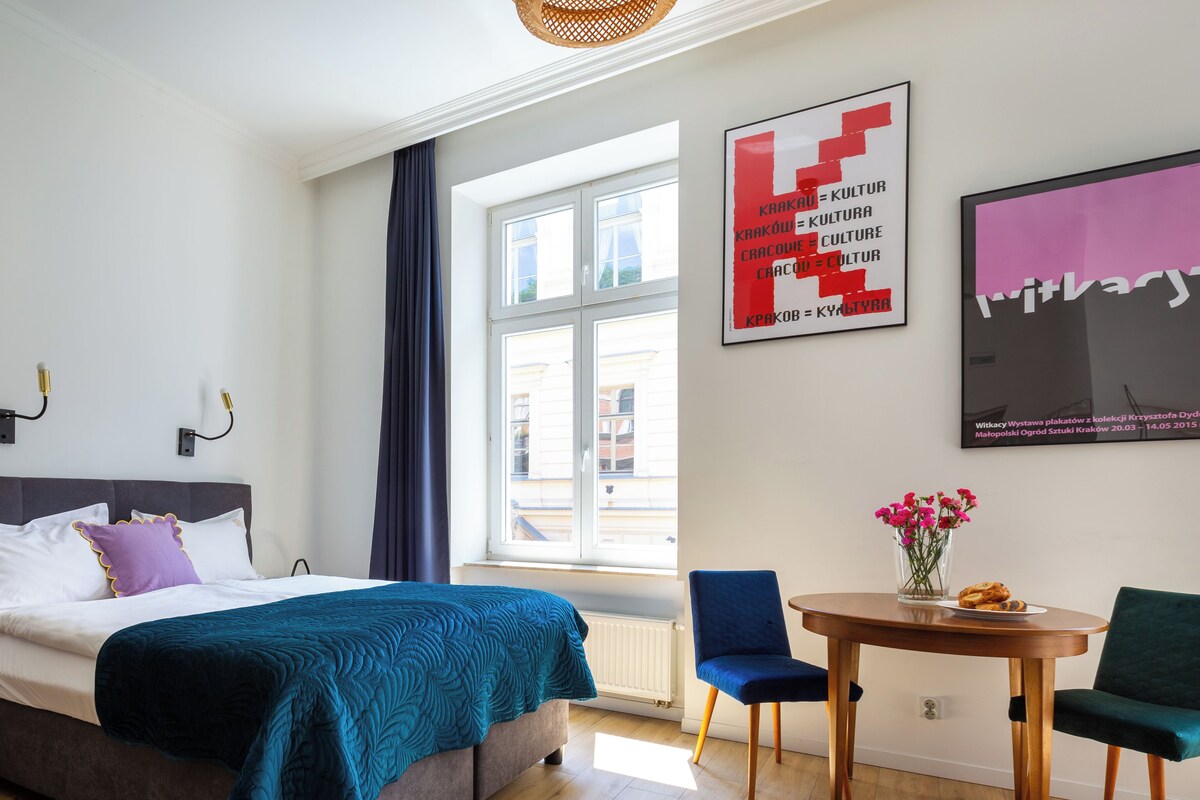 Quarto moderno com uma cama grande, uma mesa de jantar e cadeiras. Há uma janela com cortinas, uma TV fixada na parede e um pôster.