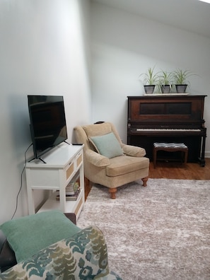 A living room with a television on a white stand, a beige armchair with a green cushion, a wooden piano, and a small table with magazines.