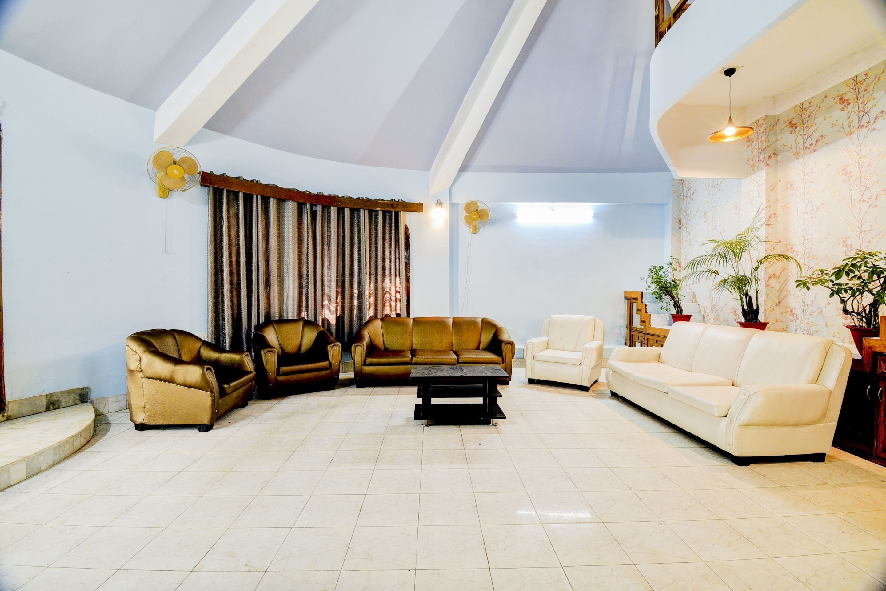 Een ruime woonkamer met een hoge plafond, een groot raam met gordijnen, en diverse zitplaatsen, waaronder banken en fauteuils.