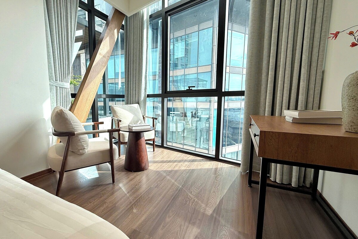 Ein Hotelzimmer mit Bett, zwei Stühlen, einem kleinen Tisch und großen Fenstern mit Blick auf die Stadt.
