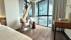 Ein Hotelzimmer mit Bett, zwei Stühlen, einem kleinen Tisch und großen Fenstern mit Blick auf die Stadt.