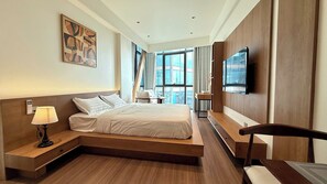 Ein Hotelzimmer mit Bett, Nachttisch, Lampe, Fernseher und Blick auf die Stadt durch große Fenster.