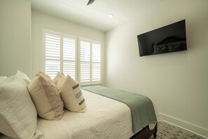 A bedroom with a bed, pillows, a green blanket, a wall-mounted TV, and a window with blinds.