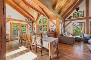 A spacious room with a wooden dining table, chairs, and a sofa. There are large windows allowing natural light and views of greenery outside. - Lavish Sanctuary Main House-Hot Tub Copake Lake (Craryville)
