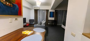 A hotel room with a wooden dining table, two grey round tables, a grey sofa, a grey armchair, a small table with a red flower, and a painting on the wall. - Terrace Studio, Costanera Views, Los Leones Metro (Providencia)