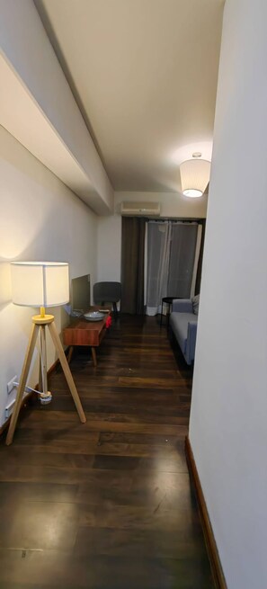 Ein schmaler Flur mit Holzboden, einer Lampe links, einem kleinen Tisch mit Teller, einem Stuhl und einem Bett mit Kopfteil rechts.