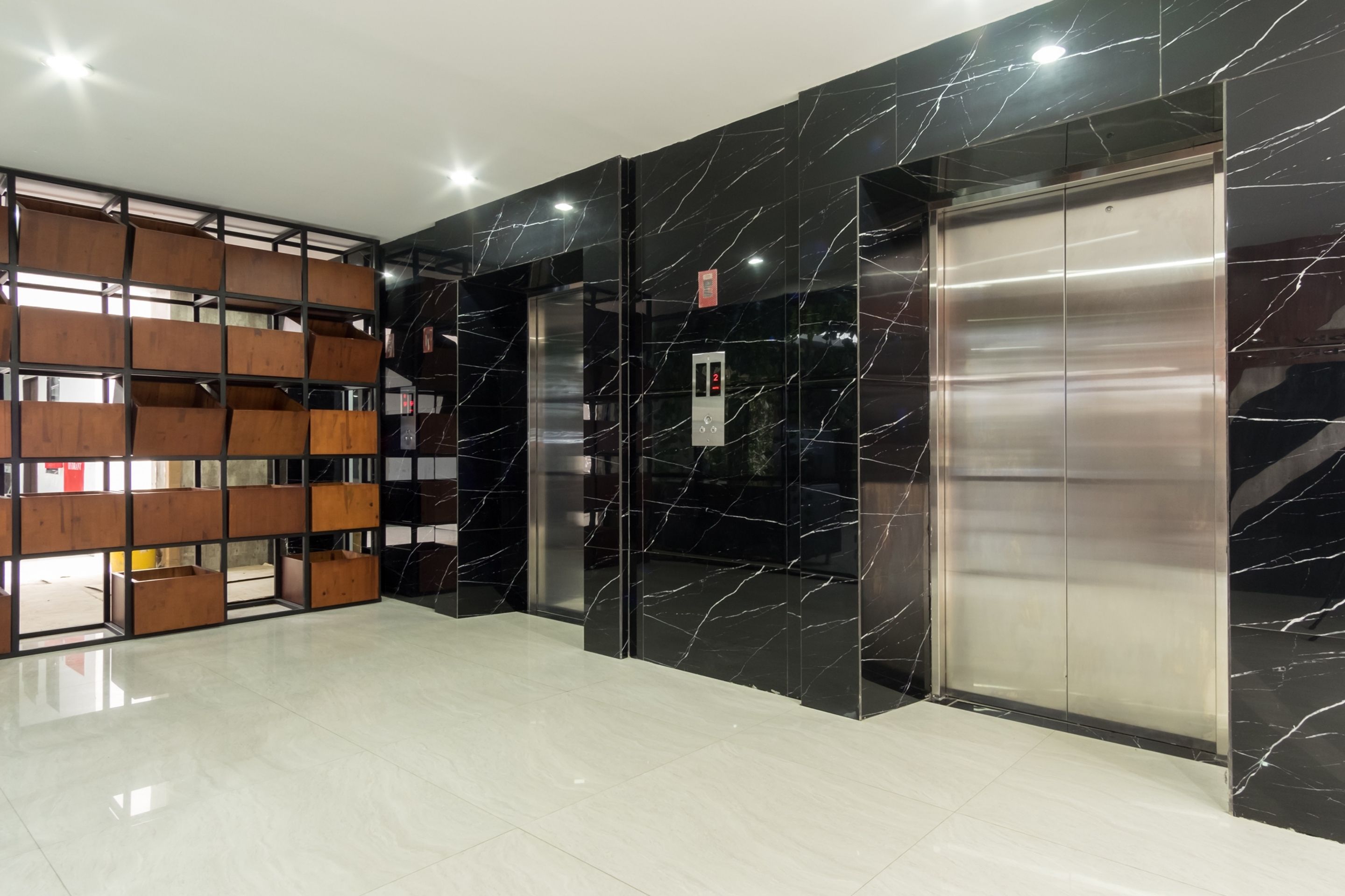 Un ascensor moderno con detalles metálicos, junto a una pared de mármol negro con vetas blancas, y una estantería de madera con varios compartimentos.