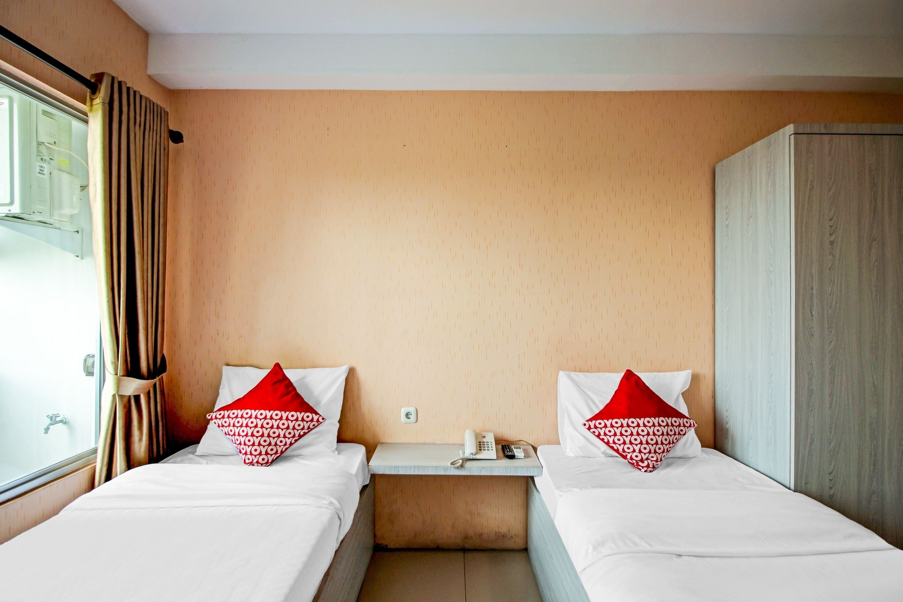 A hotel room with two beds, each with white bedding and red pillows with a repeated pattern. There is a wooden wardrobe to the right and a small bedside table with a phone and a remote control.