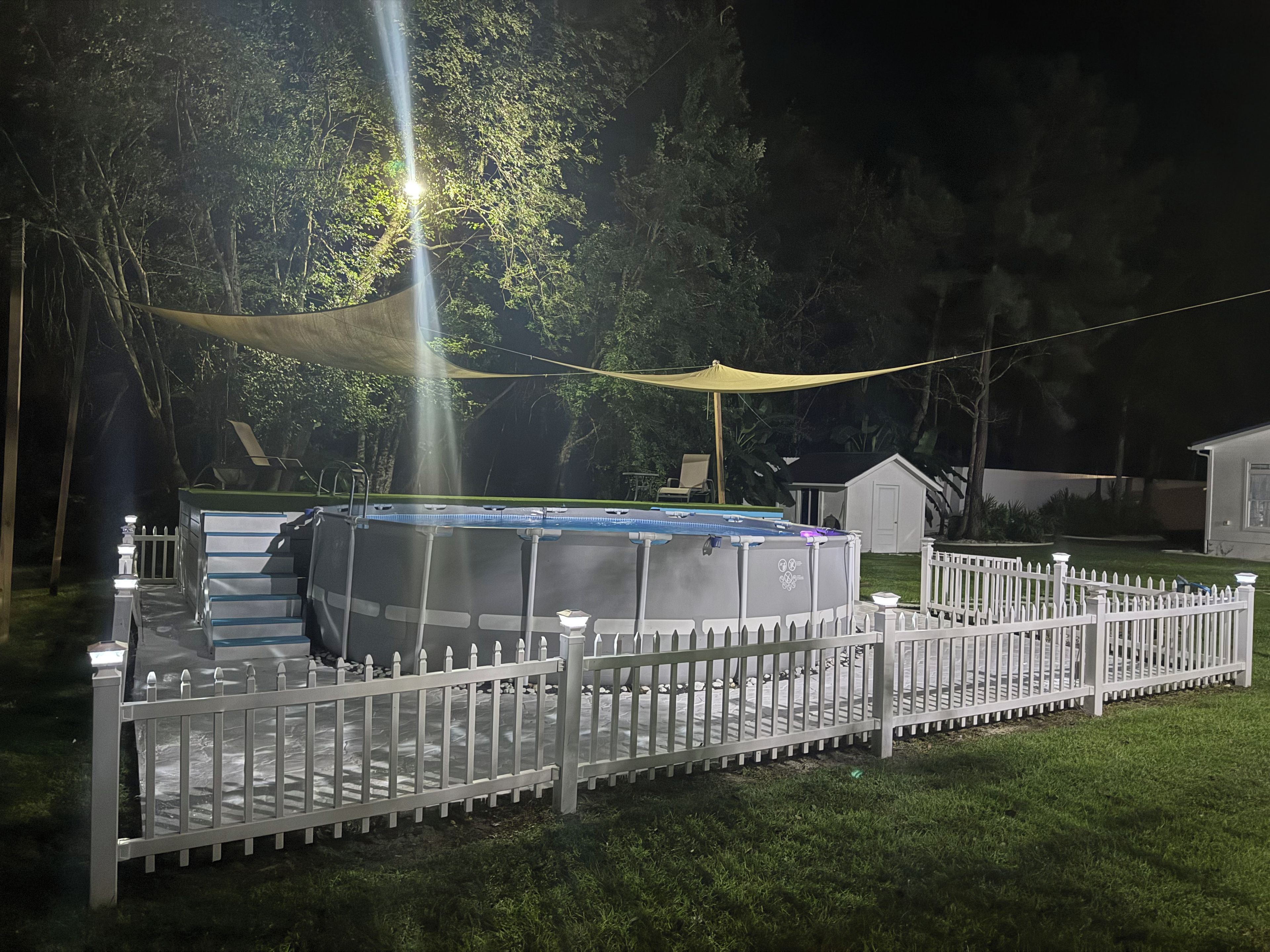 A swimming pool surrounded by a white fence, with a canopy overhead and trees in the background.